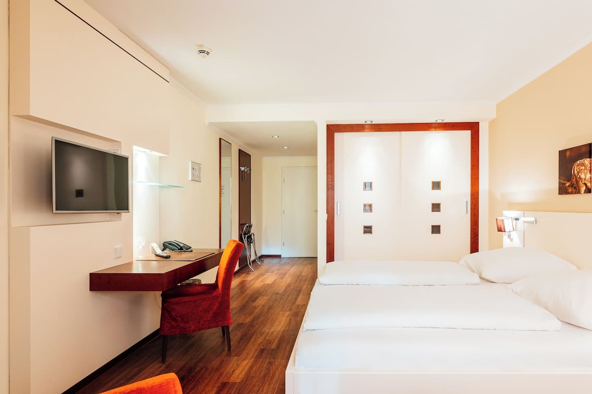 Ein Hotelzimmer mit einem Bett, einem Schreibtisch mit Stuhl, einem Fernseher und einer Tür zu einem anderen Raum.