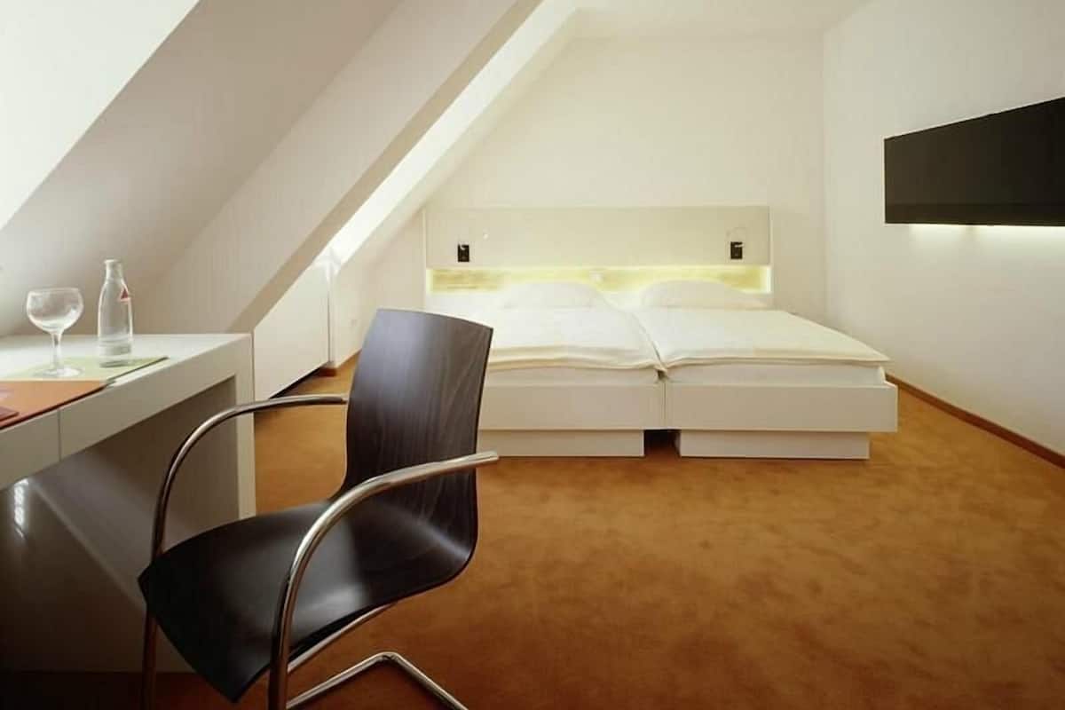 Ein modernes, minimalistisches Schlafzimmer mit schrägem Dach, einem Bett mit Kopfteil, einem Schreibtisch mit Stuhl und einem Nachttisch mit Glas und Flasche.