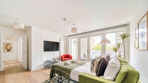 Une chambre moderne avec un canapé vert, un fauteuil rouge, un téléviseur à écran plat et une grande fenêtre donnant sur des immeubles et des arbres.