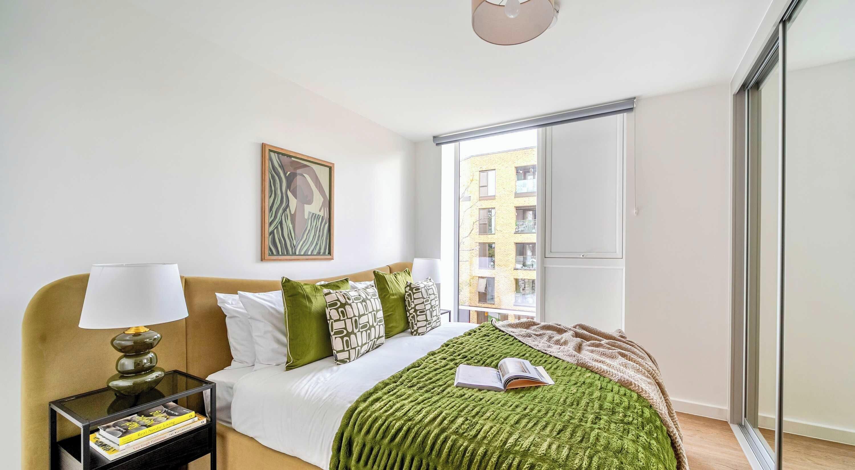 Une chambre moderne avec un lit, une table de chevet, une lampe, un tableau encadré et une vue sur les immeubles par la fenêtre.
