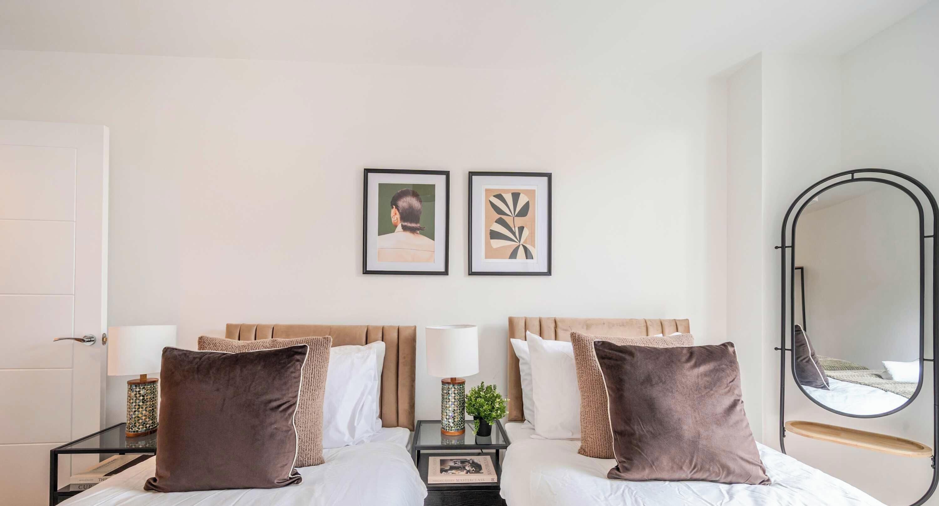 A hotel room with two beds, a bedside table with a lamp, a small plant, and a mirror on the wall.