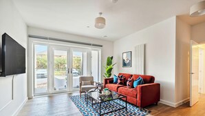 A modern living room with a large flat-screen TV, a red sofa with patterned cushions, a glass coffee table, and a large plant.