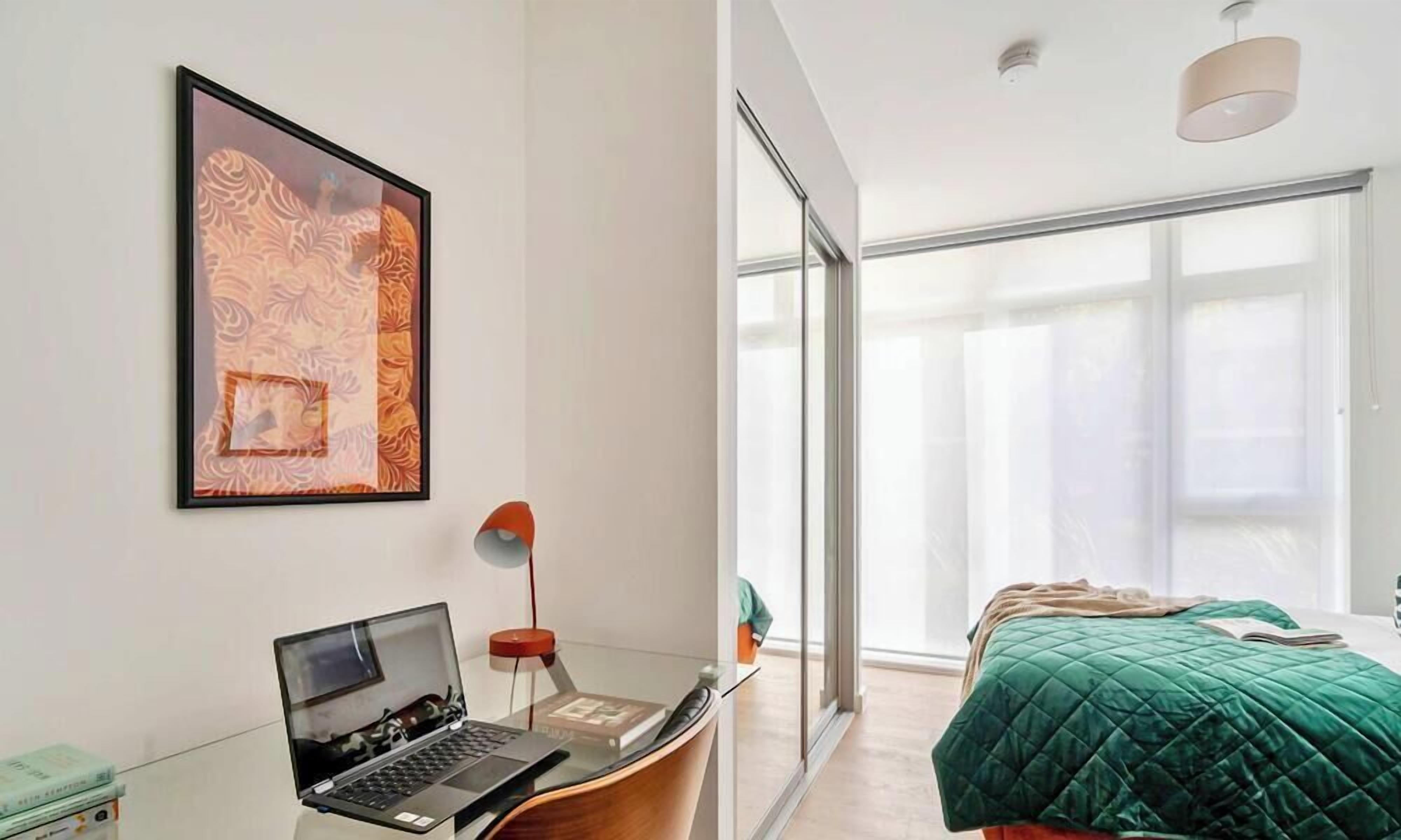 A modern bedroom with a laptop on a desk, a large window, and a green bedspread.