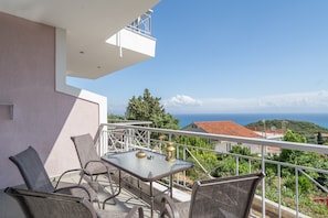 Ein Balkon mit Tisch und Stühlen, der auf eine Küstenlandschaft mit Häusern und Grünflächen blickt.