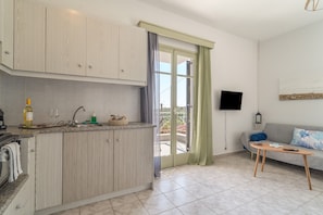Eine moderne Küche mit Einbauofen, Spüle und Arbeitsplatte mit verschiedenen Gegenständen. Eine Schiebeglastür führt zu einem Balkon mit Blick auf Grünflächen. Ein Flachbildfernseher ist an der Wand montiert, und im Wohnbereich befindet sich eine Couch mit einem Zierkissen.