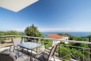En balkon med et bord og stole, med udsigt over et kystlandskab med træer og hav.