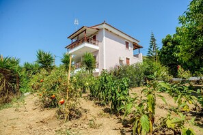 A two-story house with a balcony, surrounded by a garden with various plants and a clear blue sky.