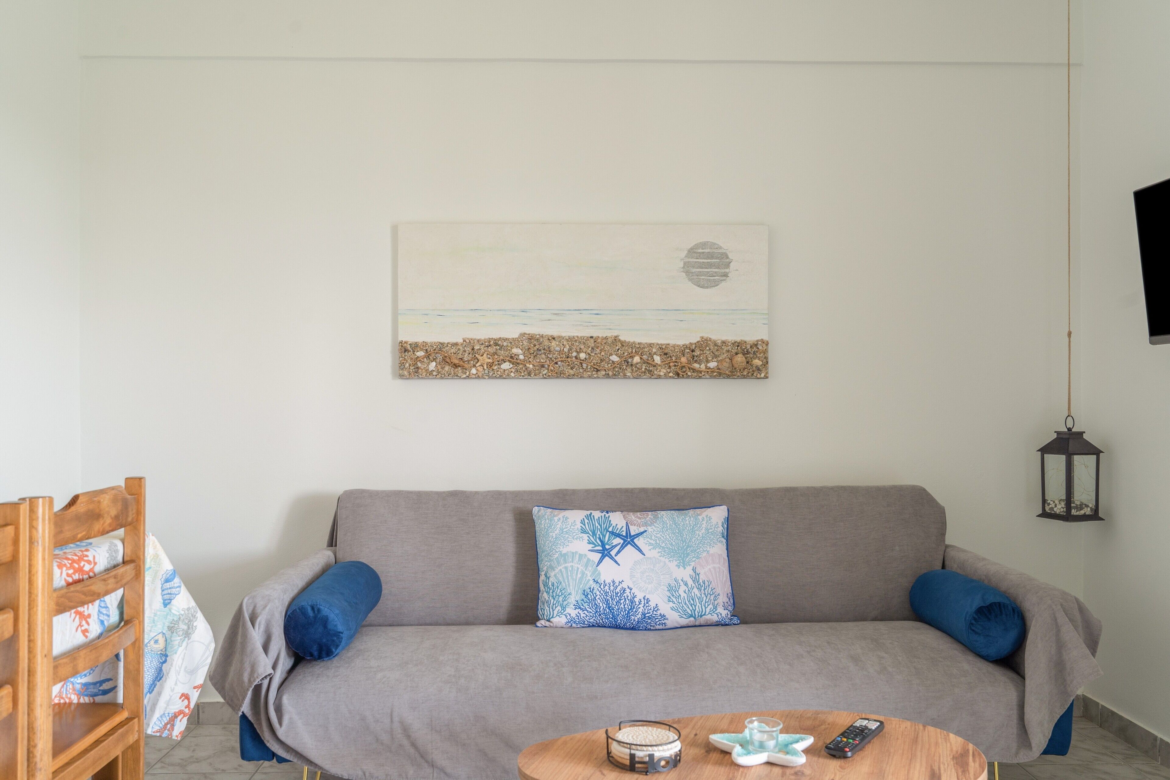 A living room with a gray sofa, a wooden chair, a round coffee table with a remote control, and a wall-mounted artwork depicting a beach scene.