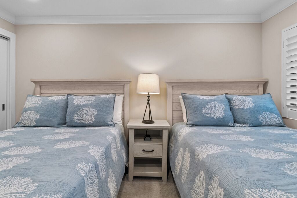 Een slaapkamer met twee bedden, elk voorzien van bijpassend blauw beddengoed en witte koraalmotieven. Er staat een nachtkastje met een lamp tussen de bedden.