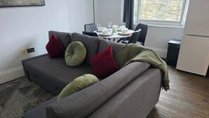 A modern living room with a grey sofa, red and green cushions, a dining table set for two, and a view through a window. - Apartment - Sleeps 6 & Near Station (Bedford)