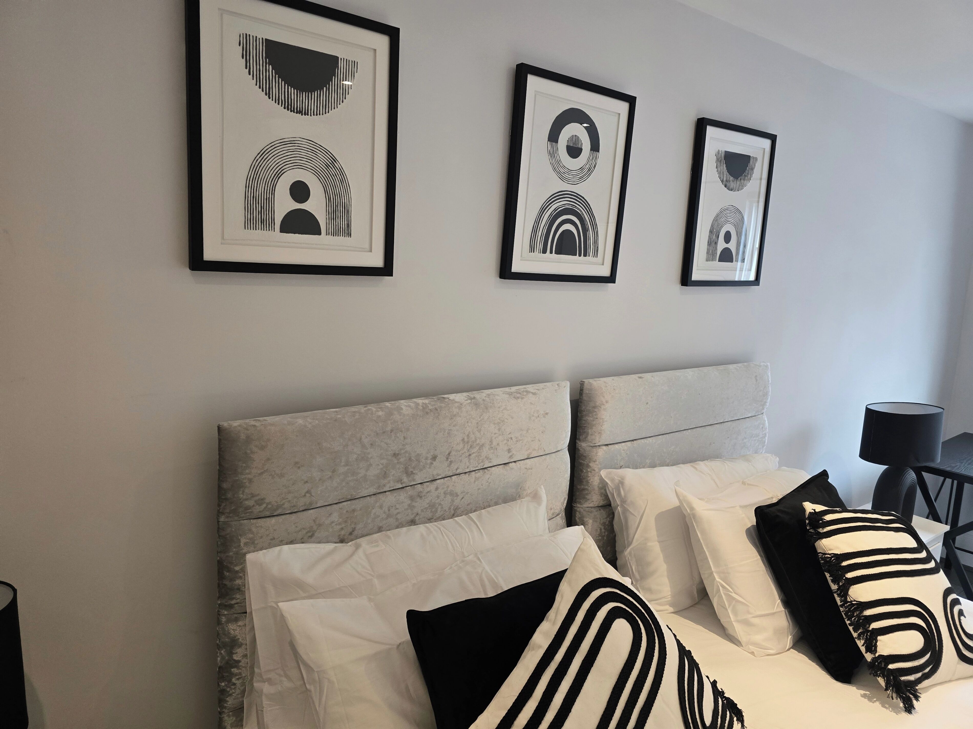 A hotel room with a bed, white and black pillows, a grey headboard, and three framed abstract artworks on the wall.