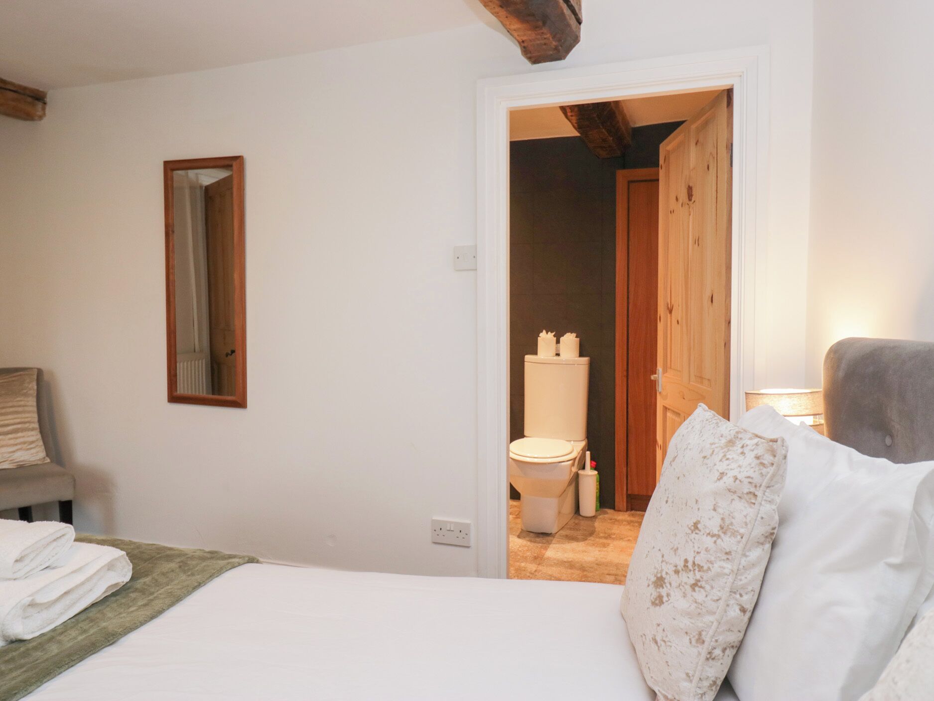 A bedroom with a bed, a bedside table with a lamp, a mirror on the wall, and a door leading to a bathroom.