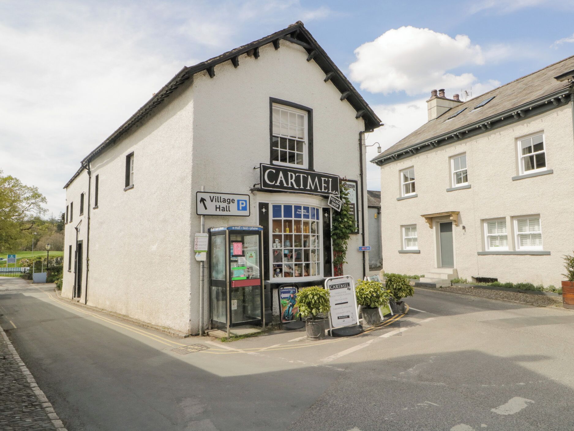 A white building with a sign reading "CARTMEL" and a Village Hall sign pointing to the left.
