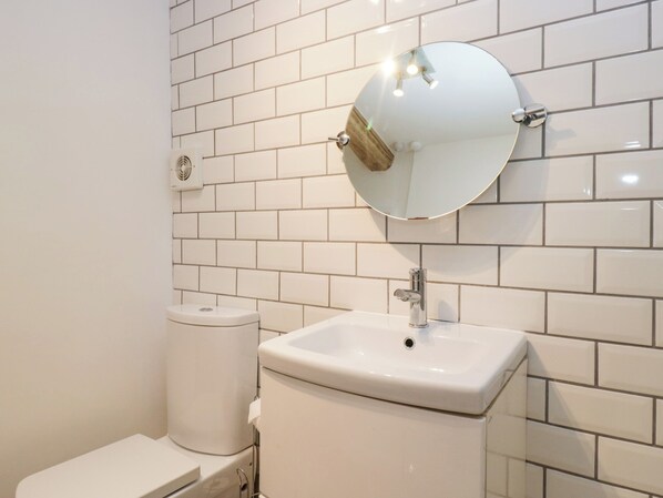 A bathroom with white tiled walls, a round mirror, a sink, and a toilet. - No.2 The Flags (Grange-over-Sands)