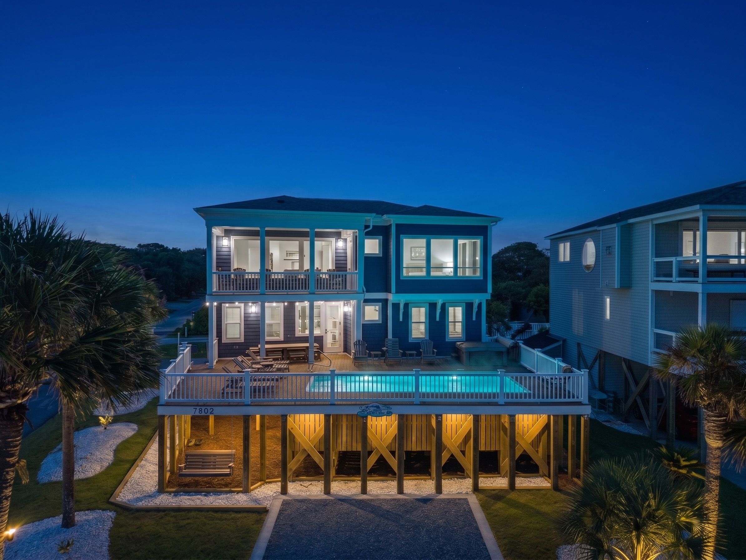 A two-story house with a pool on the ground floor, surrounded by palm trees and a clear sky.