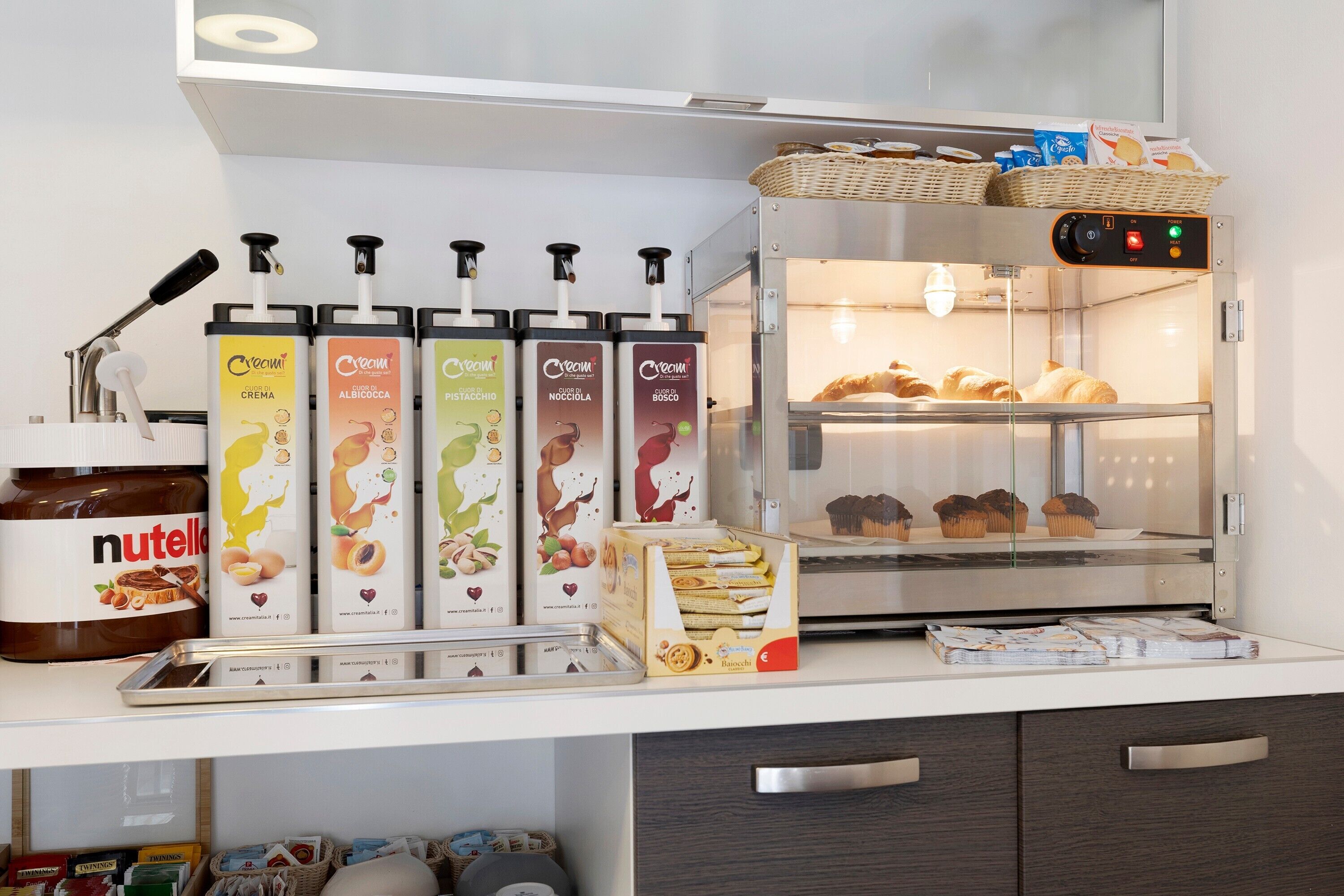 A kitchen area with a Nutella dispenser, various creamer dispensers, a bread display case, and a bread box.