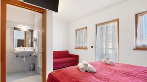 A hotel room with a red bed, a red sofa, a bathroom with a sink and mirror, and two windows with curtains.