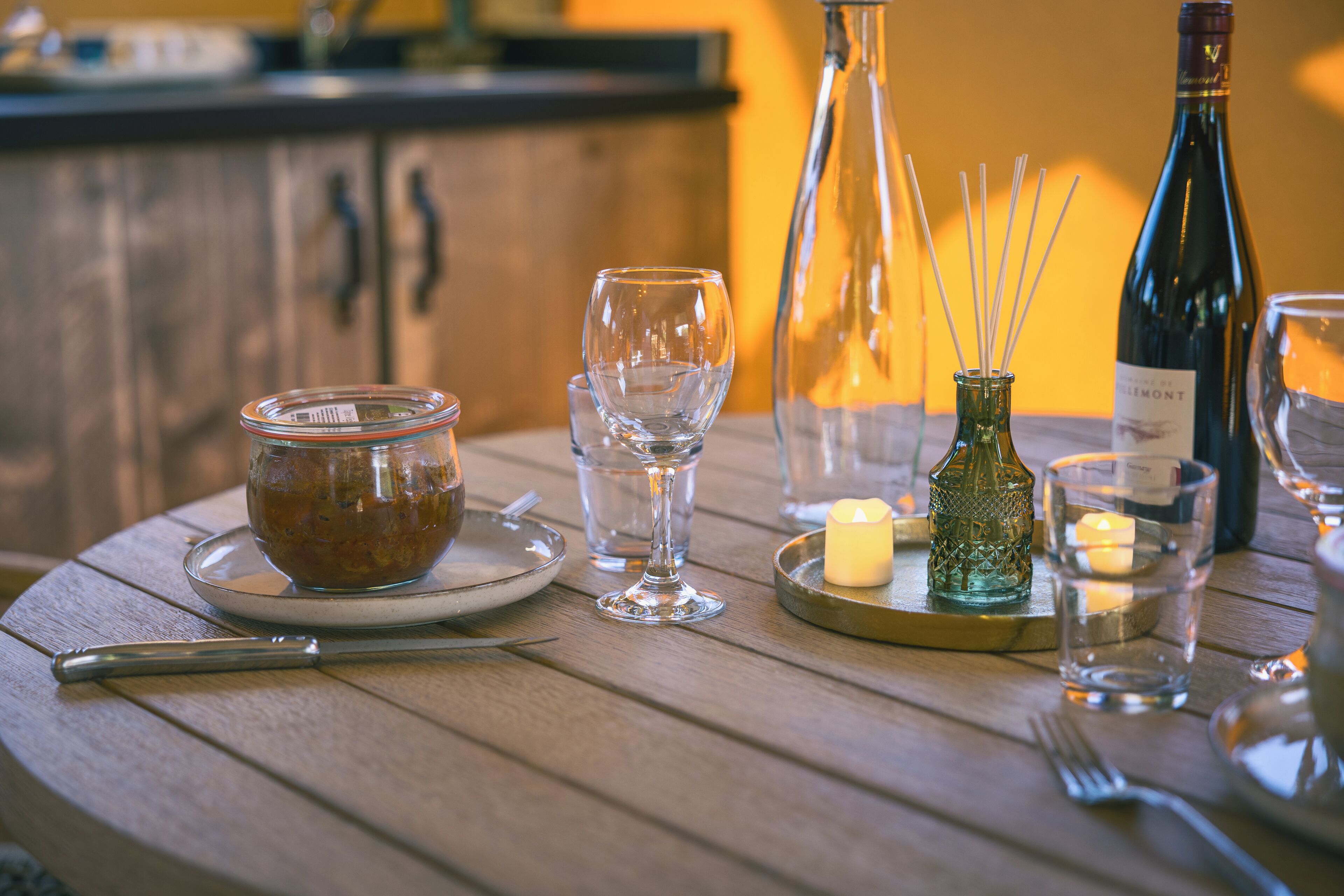 A wooden table with a glass jar, wine glasses, a candle, and a bottle of wine.
