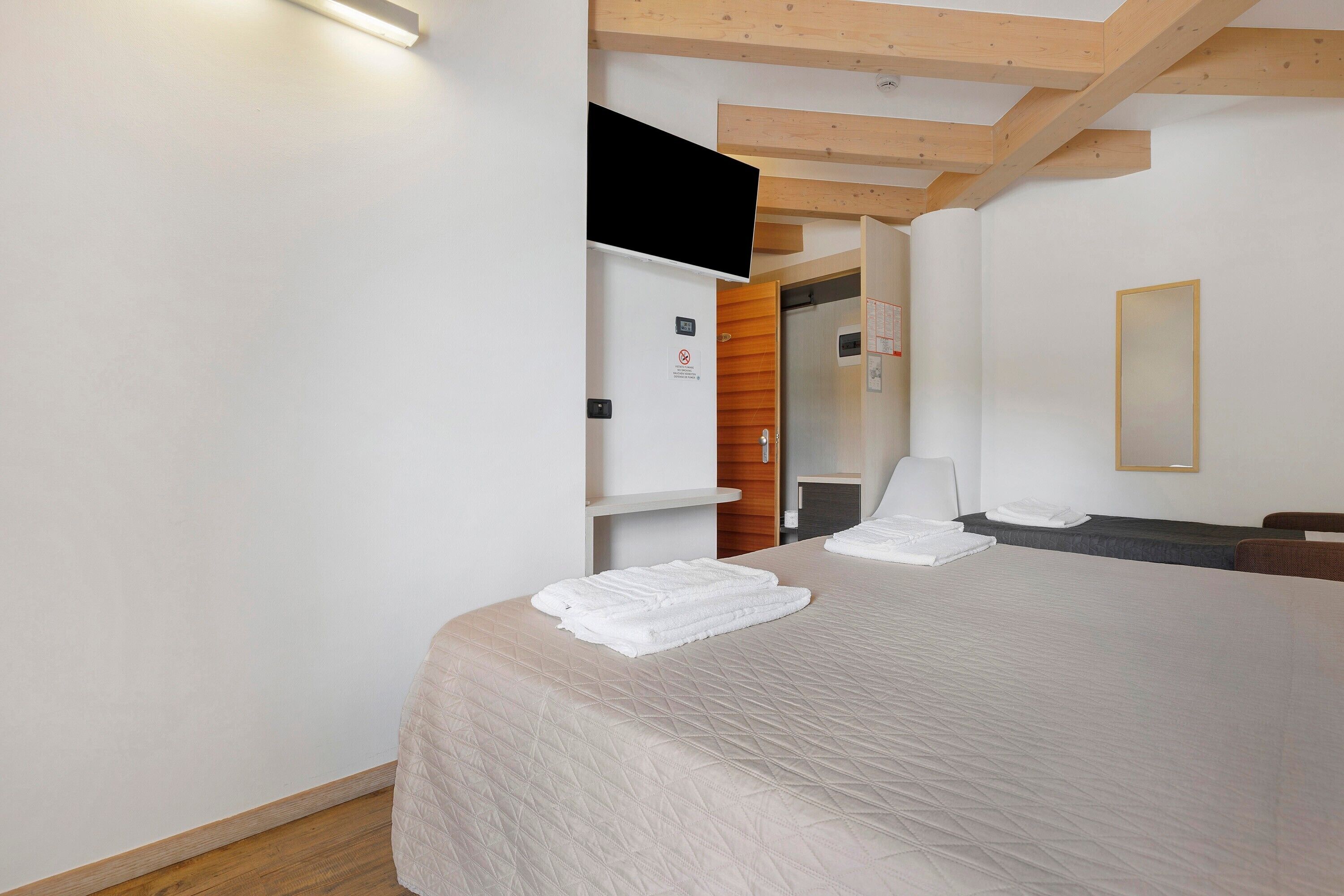 Hotellihuone, jossa on sänky, seinälle asennettu taulutelevisio, puinen katto ja ovi, joka johtaa kylpyhuoneeseen.