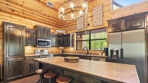 A modern kitchen with dark wooden cabinets, stainless steel appliances, a large island with stools, and a hanging light fixture. - Creekside Luxury Cabin w/ Hot Tub • Sleeps 12 (Broken Bow)