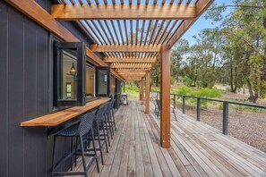 Una terrazza esterna in legno con una pergola, angolo bar con sgabelli e vista su una zona boschiva.