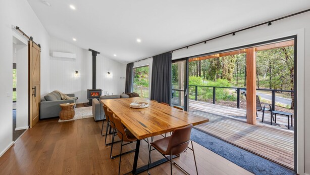 A modern interior with a wooden dining table, grey sofas, and a large sliding glass door leading to a deck with a view of a forest.