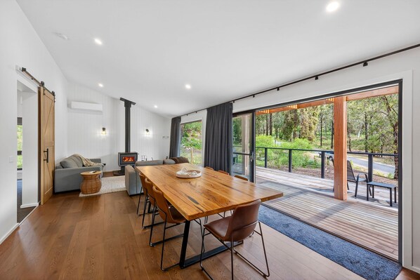 A modern interior with a wooden dining table, grey sofas, and a large sliding glass door leading to a deck with a view of a forest.