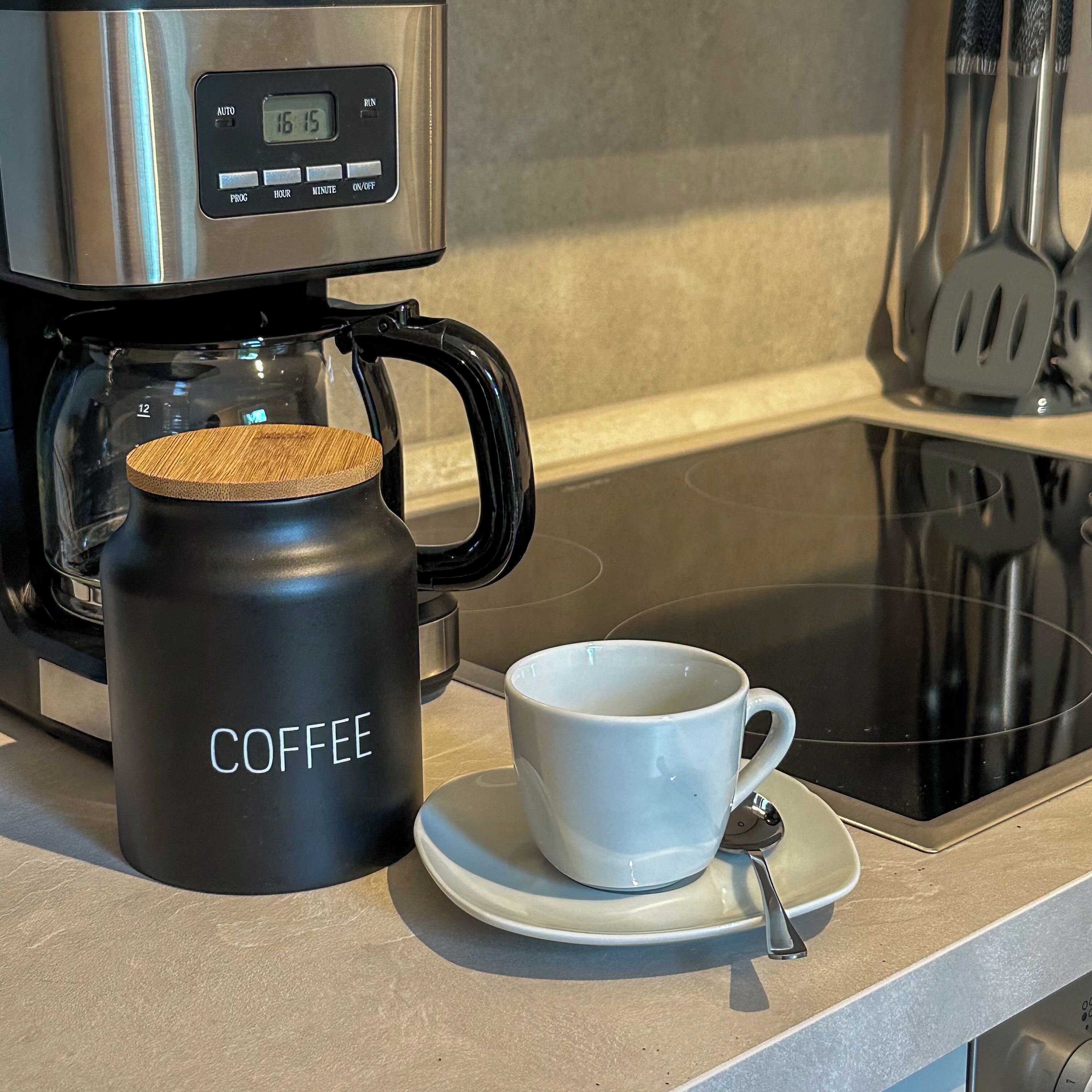 Une machine à café dotée d’un écran numérique, une tasse noire sur laquelle est inscrit le mot « CAFÉ », et une tasse blanche posée sur une soucoupe avec une cuillère.