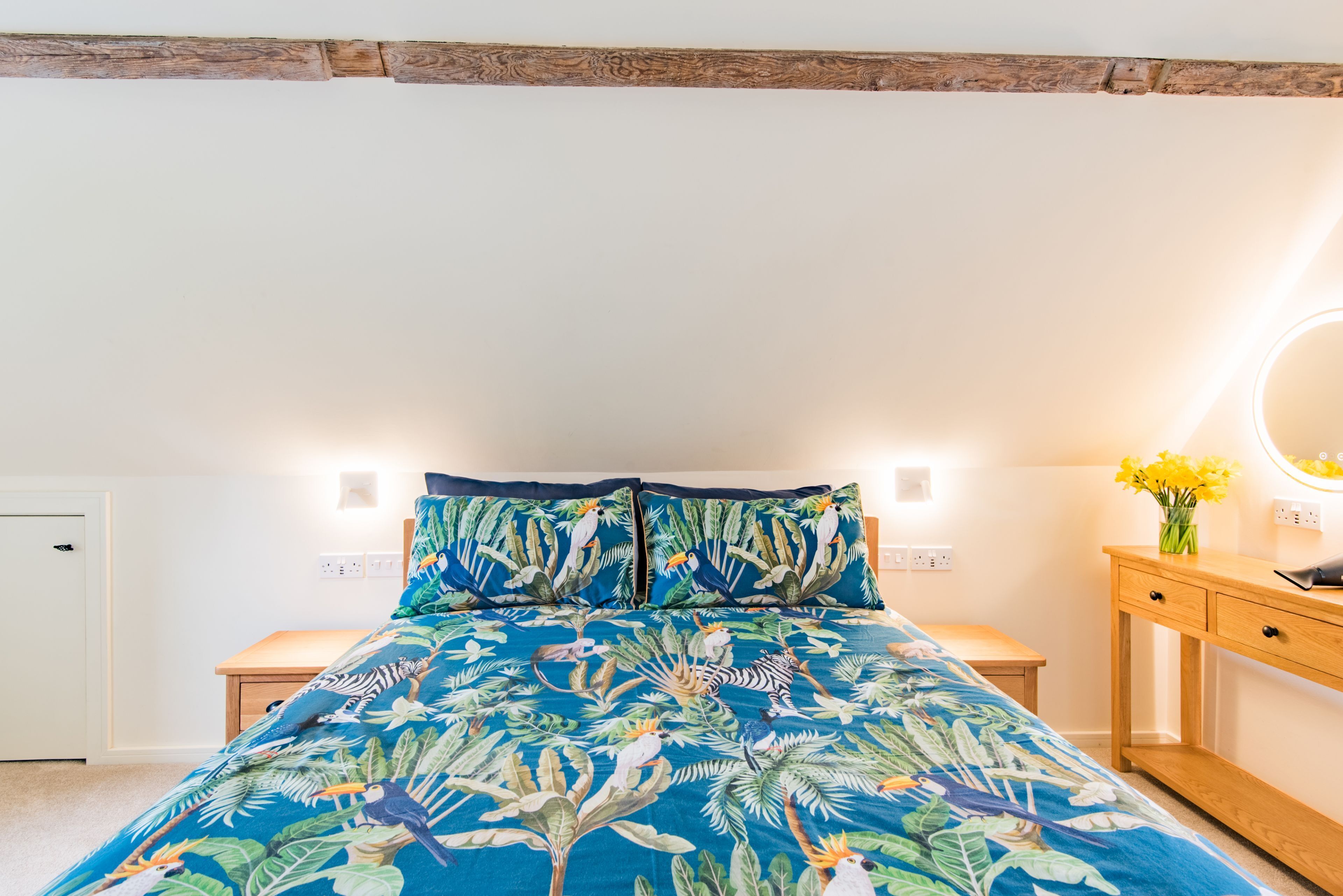 Un dormitorio con cama, mesitas de noche, cómoda de madera, espejo y un florero con flores.