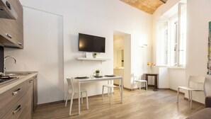 A modern kitchen with a built-in oven, a dining area with a table and chairs, a TV mounted above the counter, and a window with white shutters.