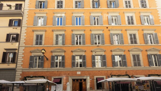 A multi-story building with a yellow facade, multiple windows with shutters, and a ground-level entrance with a sign.