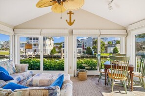 A conservatory with a ceiling fan, a cushioned sofa, a dining table with chairs, and a view of a residential area through large windows. - Coastal Retreat in South Bethany (South Bethany)