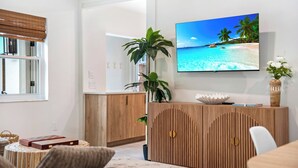 A modern living room with a wooden cabinet, a television displaying a beach scene, and a large plant. - Walk to Beach- Private Pool • Mini Golf • Firepit (Redington Beach)