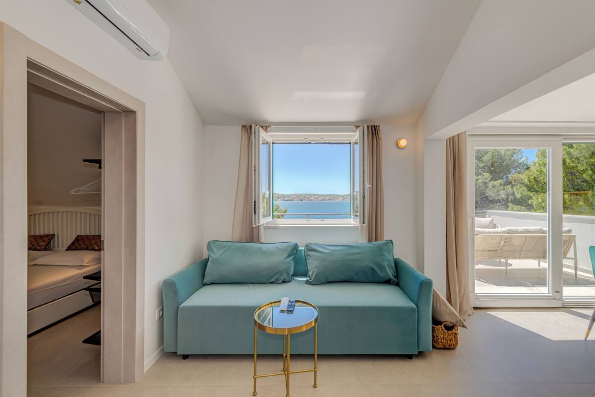 A room with a sofa, a small table, a window with a view, and a sliding door leading to a balcony.
