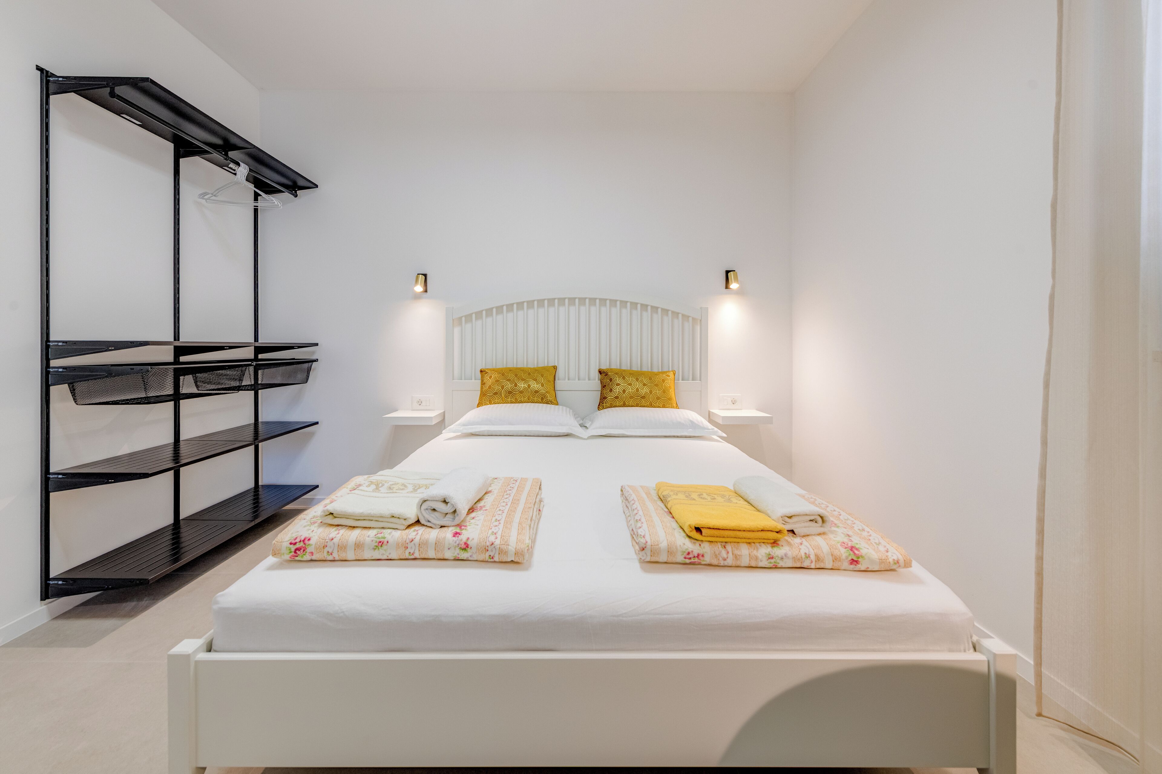 A single bed with a white headboard, two bedside tables with lamps, a black shelving unit, and a small hanging rack.