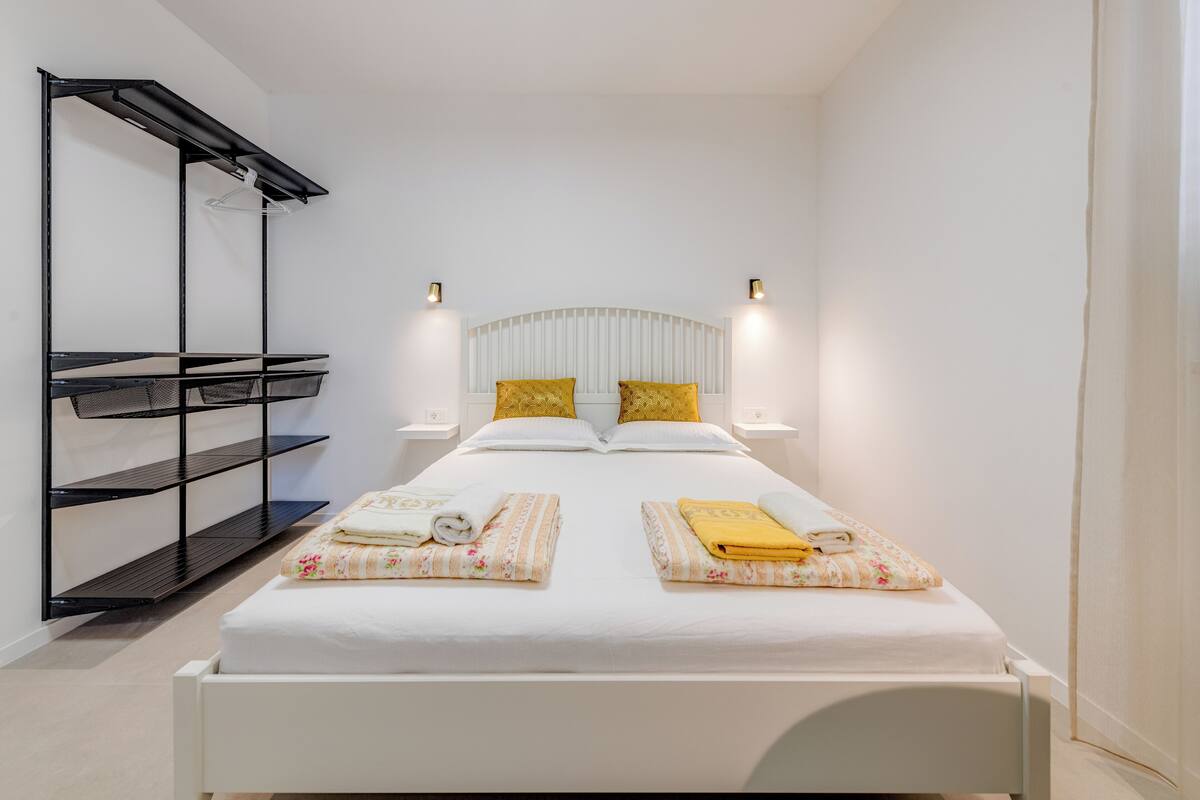 A single bed with a white headboard, two bedside tables with lamps, a black shelving unit, and a small hanging rack.