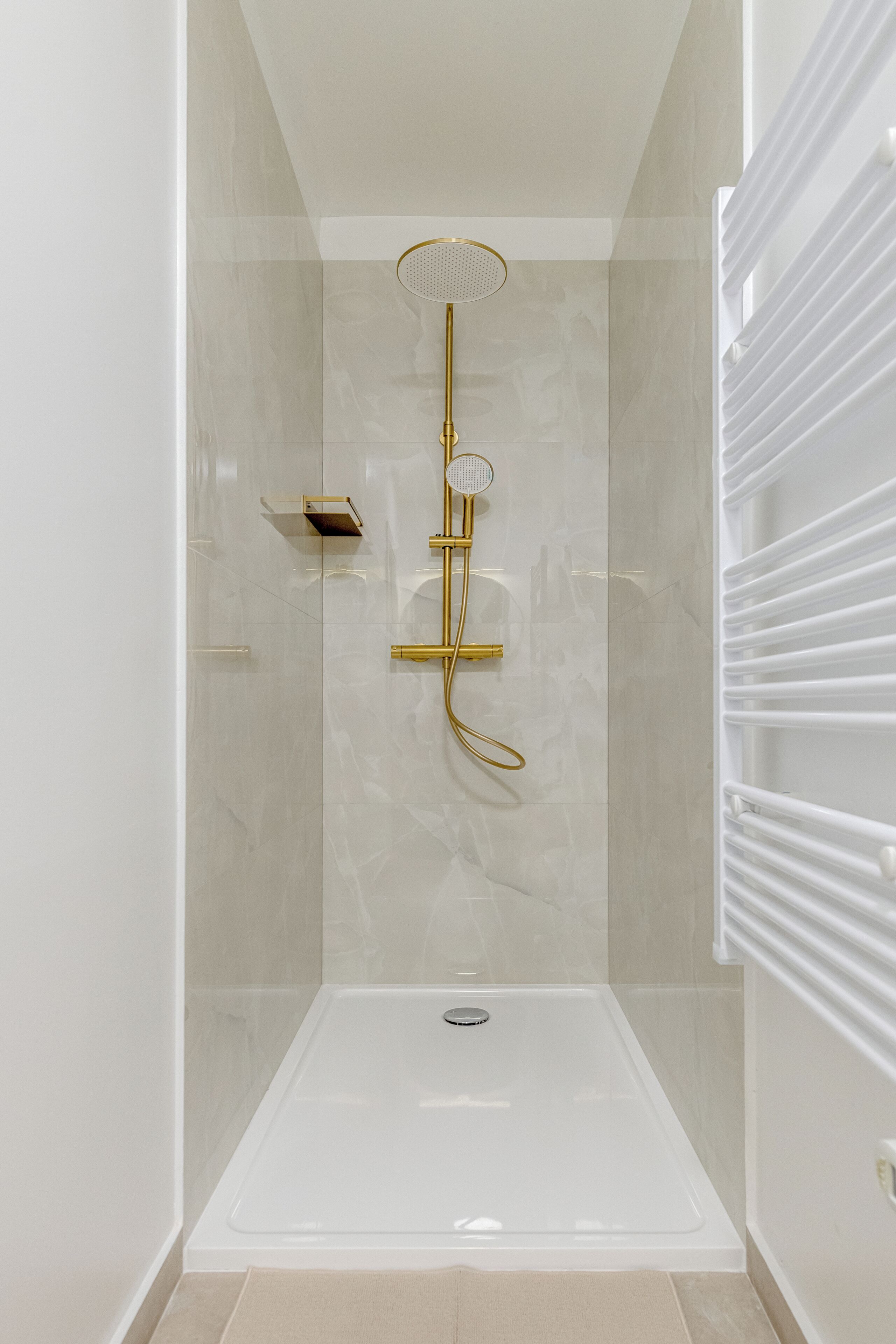 A modern shower with a gold showerhead and control panel, a white bathtub, and a beige tiled floor.