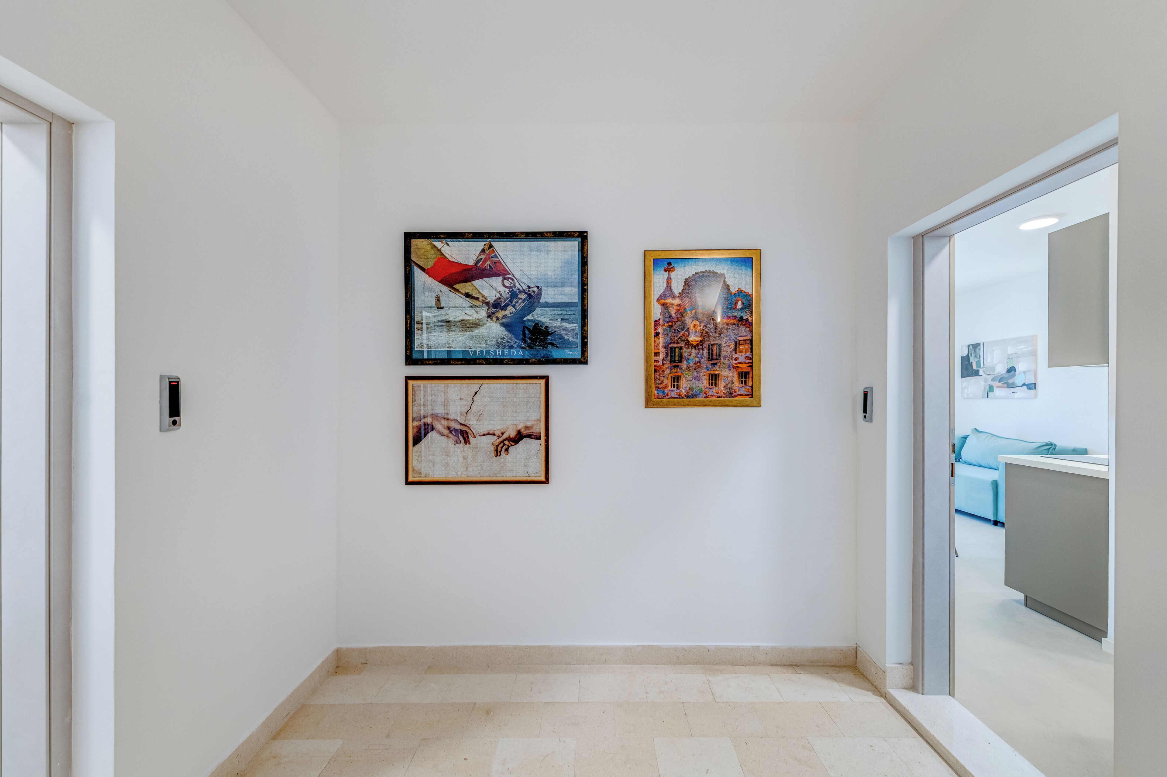 A hallway with three framed artworks on the wall, a door on the left, and a reception area visible through an open door on the right.