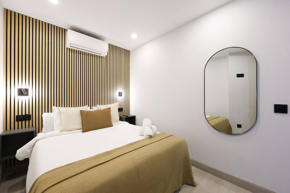 Ein modernes Hotelzimmer mit einem großen Bett, einer Wand-Klimaanlage, einem Nachttisch und einem Spiegel.