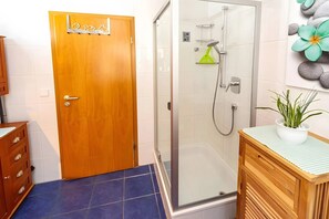 Une salle de bain avec une porte en bois, une douche en verre, un lavabo agrémenté d’une plante en pot et un sol carrelé.