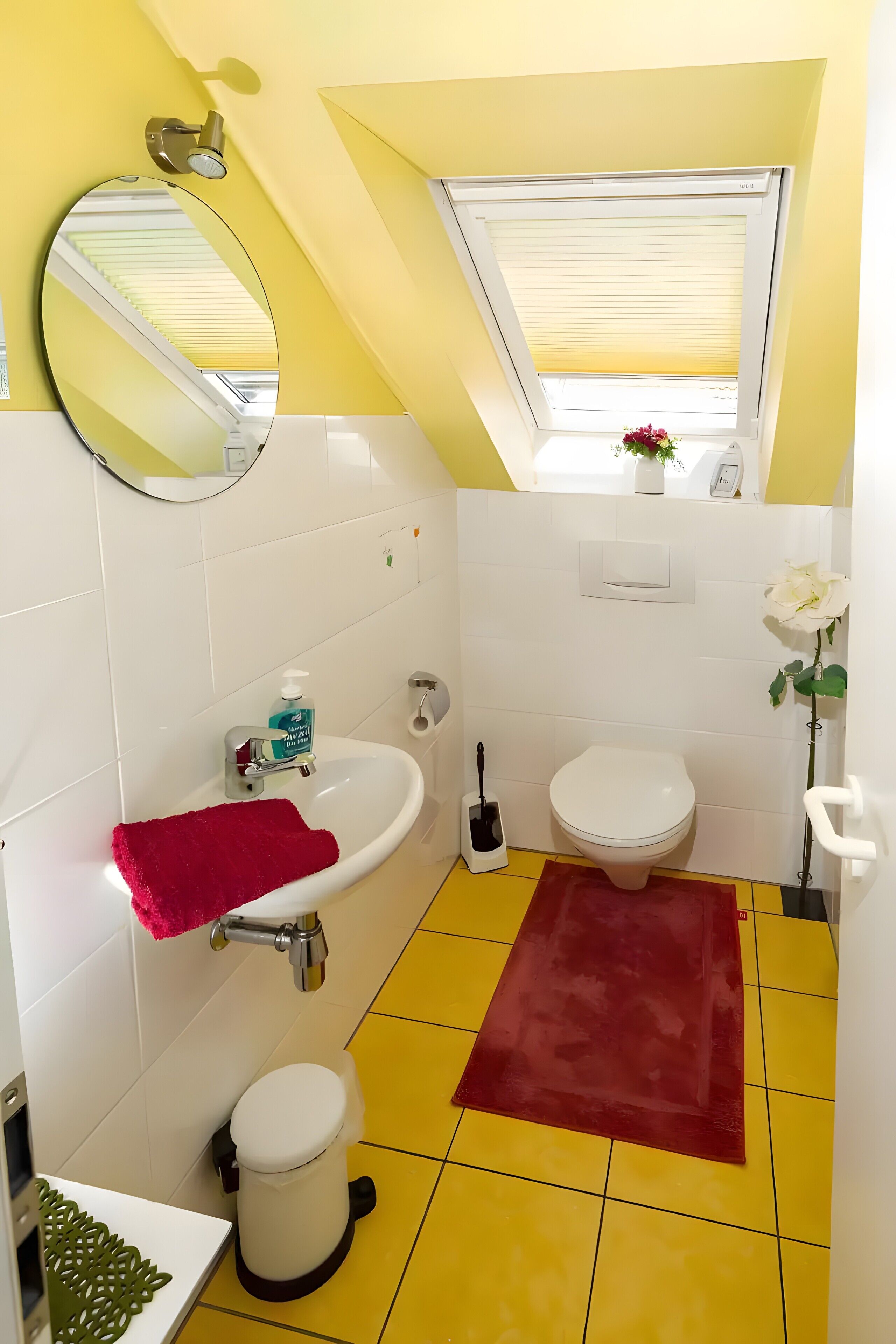 Een badkamer met een witte wastafel, toilet en bidet, een rode badmat, een spiegel en een raam met jaloezieën.