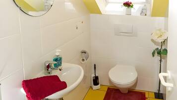 Een badkamer met een witte wastafel, toilet en bidet, een rode badmat, een spiegel en een raam met jaloezieën.