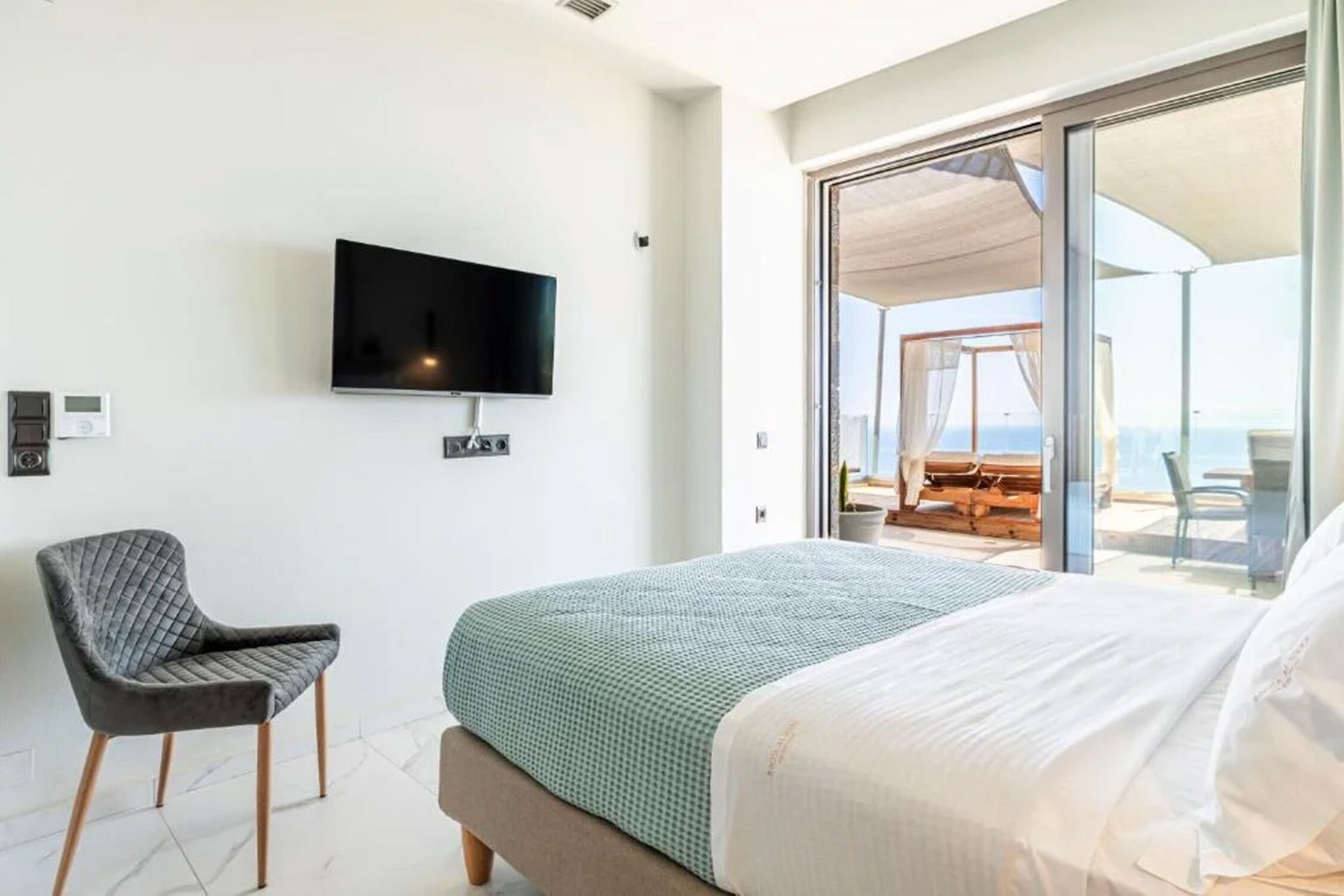 Un dormitorio con cama, una silla, un televisor y un ventanal corredero que da a un balcón con mobiliario exterior.