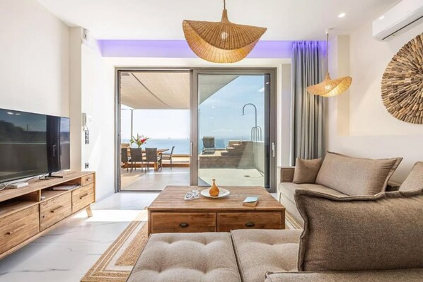 A modern living room with a large flat-screen TV, wooden furniture, and a view of the ocean through large sliding doors. - Eros Villa Crete (Malevizi)