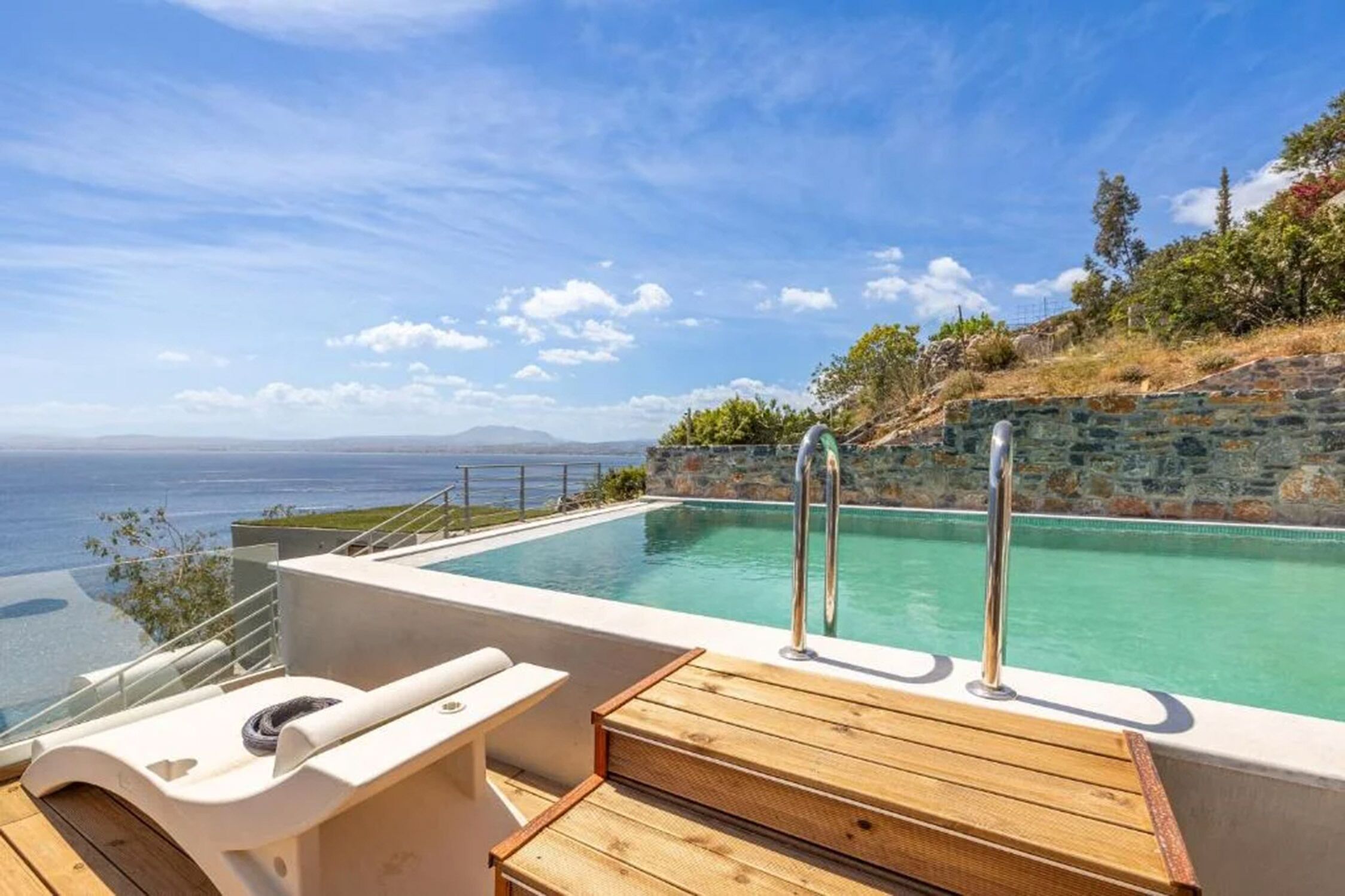 A pool with a wooden deck and metal railings, overlooking a scenic coastline.