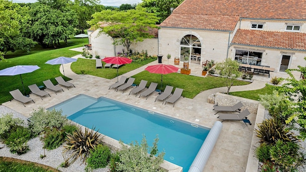 Área de piscina com espreguiçadeiras, guarda-sóis e um edifício com telhado telhado.
