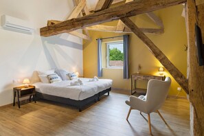 A bedroom with a bed, bedside table, chair, window, and wooden beams. - Gîte Les Lilas - Les Carassonnes with Shared Pool, Wi-Fi, and Air Conditioning (Saint-Nexans)