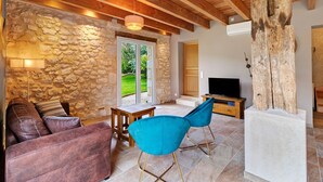 Un salon avec un mur en pierre, des poutres en bois au plafond, une cheminée en pierre, un téléviseur et un grand tronc d’arbre.