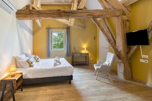 A bedroom with a bed, bedside table, lamp, chair, window, and a TV mounted on the wall. - Gîte Les Lilas - Les Carassonnes with Shared Pool, Wi-Fi, and Air Conditioning (Saint-Nexans)