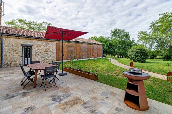 A patio with a stone table and chairs, a red umbrella, and a wooden planter with plants. - Gîte Les Lilas - Les Carassonnes with Shared Pool, Wi-Fi, and Air Conditioning (Saint-Nexans)